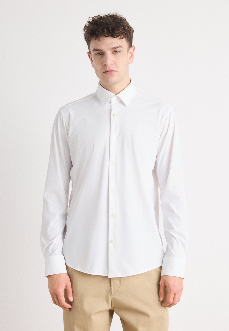 Hvit skjorte med lange ermer, spredt krage, glatt stoff og slim fit, vist innstoppet i beige bukser.