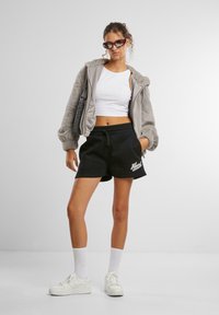 Flauschiger grauer Reißverschluss-Hoodie über einem weißen kurzen Tanktop, kombiniert mit schwarzen Shorts mit weißem Logo und weißen Sneakers mit strukturierten Schnürsenkeln.