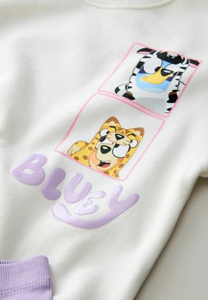 Biela mikina s dvoma farebnými zvieracími grafikami, ktoré zobrazujú zebru a leopardu, a slovom „BLUEY“ v purpurových, lesklých písmenách.