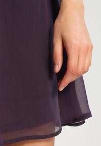 Paarse doorschijnende stof met een textuurafwerking. De zoom is licht gebogen en een hand raakt voorzichtig de stof aan.