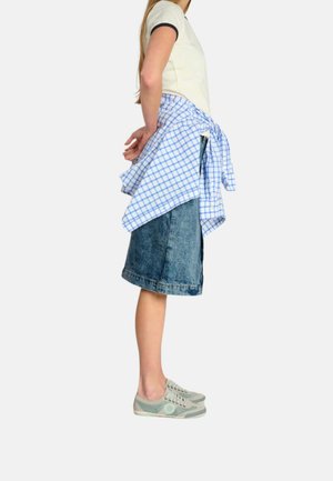 Mujer con camiseta blanca, falda vaquera azul hasta la rodilla, zapatillas grises, con camisa azul y blanca de cuadros atada alrededor de la cintura, de pie de lado.