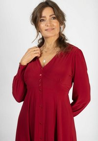 Punainen pitkähihaine mekko, jossa on V-aukko, nappi edessä ja rypytettyjä yksityiskohtia rinnoissa. Tasainen pinta ja pehmeä kangas.