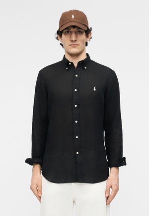 Vyras vilki rudą kepuraitę ir juodą ilgomis rankovėmis marškinius su baltu išsiuvinėtu logotipu, stovi priešais šviesią paprastą foną.