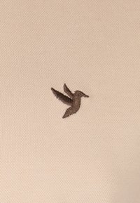Beige stof med en detaljeret brun broderet kolibri, der viser vingerne i midt-vingeslag, og en blød, tekstureret overflade.