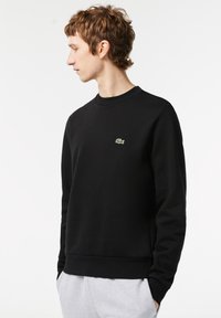 Svart sweatshirt med rundad halsringning, ribbade ärmslut och nederkant; har en liten grön Lacoste-logotyp på bröstet; slät textur.
