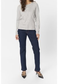 Frau, die eine hellgraue, geknöpfte Strickjacke, dunkelblaue Jeans und schwarze spitze High Heels mit drei Riemen und goldenen Schnallen trägt.