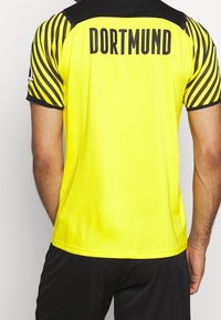 Gul sporttröja med svarta detaljer och randiga ärmar. Har texten "DORTMUND" tydligt på ryggen. Lätt och ventilerande tyg.