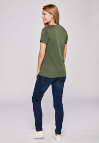 Oliveově zelené bavlněné tričko s krátkými rukávy, volný střih, kulatý výstřih, kombinované s tmavě modrými slim-fit džínami a bílými teniskami.