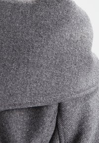 Tejido gris de punto con un patrón texturizado, que presenta un cuello doblado y un diseño que cae de forma suelta. Apariencia suave y cálida.