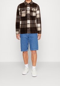 Camisa chaqueta de forro polar a cuadros marrón y crema con dos bolsillos en el pecho, combinada con pantalones cortos de mezclilla azules y zapatillas blancas.