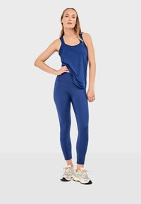 Femme debout, main sur la hanche, portant un haut bleu sans manches, des leggings assortis et des baskets blanches sur un fond gris clair uni.