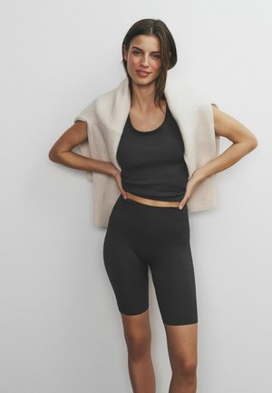 Schwarzes, geripptes Trägershirt und hoch taillierte Fahrradhosen, kombiniert mit einem hellbeigen Strickpullover, der über die Schultern gelegt ist.