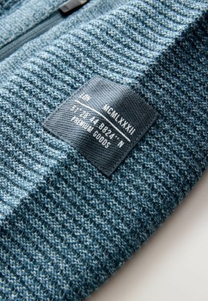 Strickpullover aus einem strukturierten, blauen Stoff mit einem schwarzen Etikett, auf dem weiße Schrift zu sehen ist. Das Design umfasst Streifen und einen Reißverschluss.