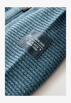 Gebreide trui in een gestructureerde, blauwe stof met een zwart etiket waarin witte tekst staat. Het ontwerp bevat strepen en een rits.