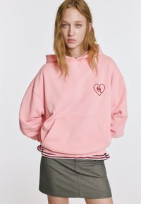 Ružová mikina s kapucňou s predným vreckom, srdcovým logom na hrudi a rebrovaným lemom. Nosená cez sivú sukňu s jemnou textúrou a prispôsobeným strihom.