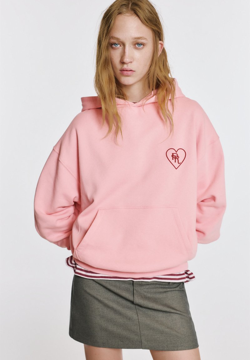 Ružová mikina s kapucňou s predným vreckom, srdcovým logom na hrudi a rebrovaným lemom. Nosená cez sivú sukňu s jemnou textúrou a prispôsobeným strihom.