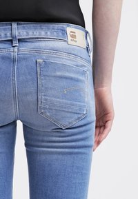 Ljust blå denimjeans har en figurnära design med subtila blekningseffekter, en bakficka med sömnadsdetalj och en beige etikett nära midjan.