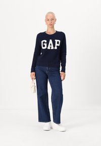 Marineblauwe trui met witte "GAP" letters, gecombineerd met wijdvallende blauwe jeans en witte sneakers. Model houdt een kleine crèmekleurige tas vast, minimale achtergrond.