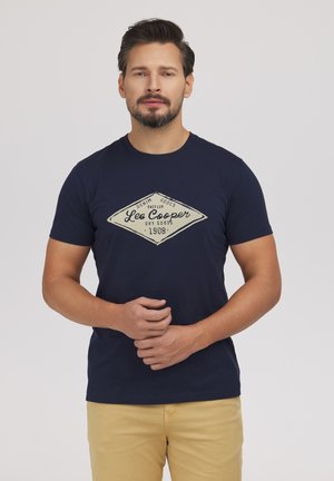 Mężczyzna w granatowym t-shircie Lee Cooper i beżowych spodniach, stoi ze splecionymi rękami z przodu na jednolitym szarym tle.