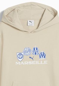 Beżowa bluza z kapturem z logo drużyny sportowej Marsylii oraz słowem "MARSEILLE" w kolorze białym, z logo Pumy w kolorze niebieskim.