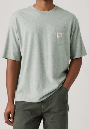 Mężczyzna noszący jasnoszary T-shirt z krótkim rękawem z kieszenią na klatce piersiowej, na której znajduje się mała naszywka z czerwonym tekstem, oraz ciemnoszare spodnie.