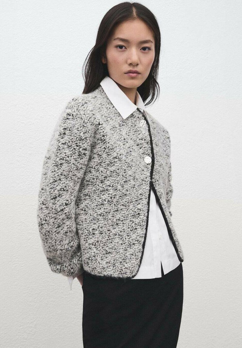 Smeđi džemper sa sivim i bijelim mrljama, okruglim ovratnikom, zatvaranjem na gumbe sprijeda te crnim ukrasnim obrubom, nošen preko bijele košulje i crne suknje.