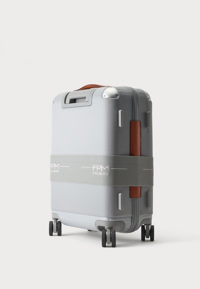 Valise rigide grise avec quatre roues, poignée en cuir marron et bande grise portant la mention FPM Milano autour du milieu.