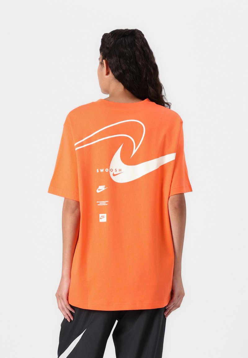 Oranžs kokvilnas T-krekls ar lielu baltu svītru grafiku un "SWOOSH" tekstu uz muguras. Izstrādāts ar brīvu piegriezumu un īsām piedurknēm.