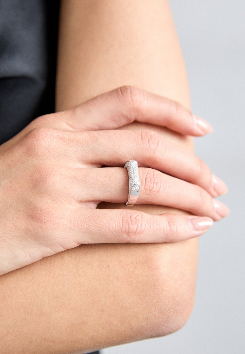 Ezüst gyűrű textúrált felülettel és egy kis gyémánt díszítéssel. Finoman manikűrözött körmű kézen viselve, semleges háttér előtt.