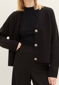 Černý texturovaný kardigan s žebrovanými manžetami a přímým střihem, zdobený stříbrnými knoflíky a nošený přes tmavý žebrovaný top.