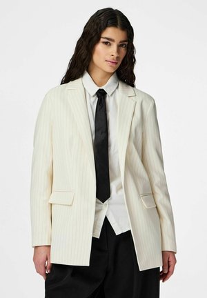 Frau mit langen dunklen Haaren, die ein weißes Nadelstreifen-Blazer, ein weißes Hemd, eine schwarze Krawatte und schwarze Hose vor einem schlichten Hintergrund trägt.