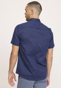 Chemise à manches courtes, de couleur bleu marine, fabriquée en tissu léger. Elle possède un col boutonné et un ourlet arrondi, avec un détail de pièce de dos unique.