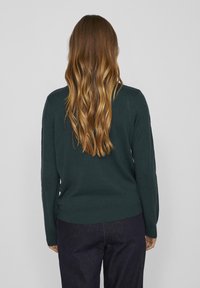 Mörkgrön tröja med långa ärmar och ribbad nederkant. Tyget verkar slätt, med en mjuk textur. Bärs med mörka, raka byxor.