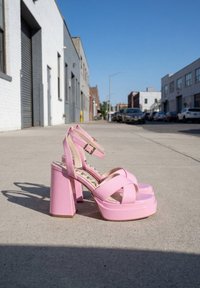 Sandales à talons hauts plateforme roses avec brides à la cheville, posées sur un trottoir en béton dans un environnement urbain.
