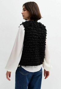 Zwarte textuurvest met een pluizig, gerimpeld ontwerp; gedragen over een wit shirt met lange mouwen. Casual uitstraling, met een cropped snit en ronde halslijn.