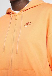 Oranssi vetoketjuhuppari, joka on valmistettu pehmeästä kankaasta ja jossa on valkoisella vuorattu huppu, kiristysnyörit ja kultainen Nike-logo rinnassa.