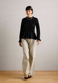 Top peplum nero con smocking testurizzato, abbinato a pantaloni larghi color avorio. Il modello indossa scarpe scure; una semplice collana di perle completa il look.