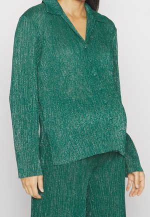 Kobieta w zielonej, długorękawowej, teksturowanej bluzce z kołnierzykiem oraz dopasowanych szerokich spodniach na tle jednolitego tła.