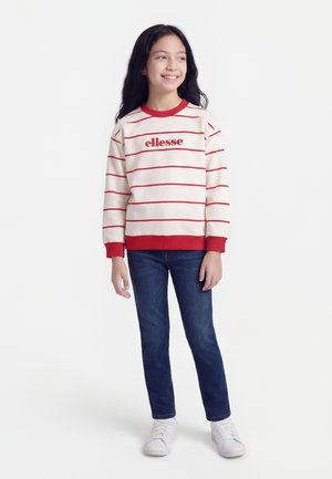 Smiling girl with long dark hair wearing a white and red striped sweatshirt, blue jeans, and white sneakers, standing on a white background.