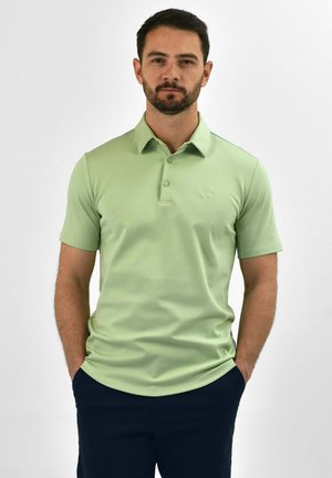 Man met donker haar en baard, gekleed in een lichtgroen poloshirt met korte mouwen en een donkere broek, staand met handen in de zakken.