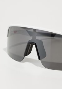Gafas de sol con un marco blanco, lentes tintados en gris oscuro, diseño envolvente y un soporte puente negro distintivo. Textura suave.