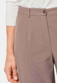 Bruine, op maat gemaakte broeken van gladde stof. Bevat een enkele knoopsluiting, vlakke voorkant en details van zijzakken.