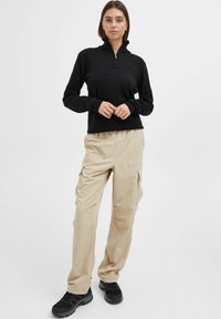 Haut en polaire noir zippé, pantalons cargo beiges avec poches latérales et chaussures noires. Texture lisse et coupe décontractée dans une palette de couleurs neutres.