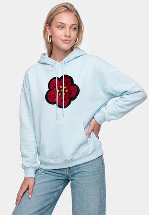 Ung kvinde iført lyseblå hættetrøje med rød og sort blomstermønster, lyseblå jeans og øreringe i ringform, stående med hånden på hoften.