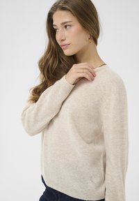 Beige, puha kötött pulóver kerek nyakkivágással, hosszú ujjakkal és bordázott mandzsettával. Finom textúrájú logó részlet található az alsó perem közelében.