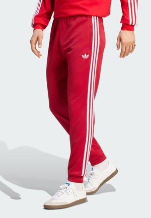 Persona con pantalones deportivos rojos de Adidas con rayas blancas y zapatillas blancas, de pie sobre un fondo claro.