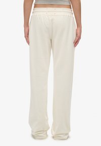 Persona vestida con pantalones de chándal sueltos de color crema con una cinturilla elástica, de pie contra un fondo blanco liso, de espaldas.