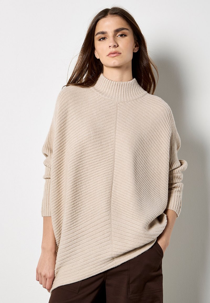 Suéter beige de cuello alto acanalado con un corte relajado y hombros caídos, que presenta un patrón texturizado y aberturas laterales en la parte inferior.