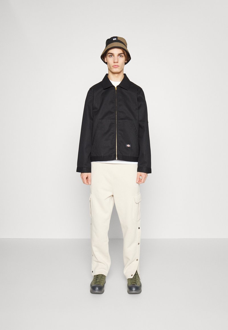 Giacca nera con zip e colletto a punta, cappello a secchiello beige, pantaloni cargo crema con bottoni a pressione e stivali neri con lacci verdi.