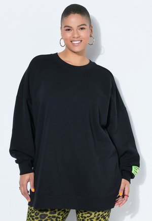 Personne souriante aux cheveux courts portant de grandes boucles d'oreilles en anneau, un sweat-shirt noir surdimensionné avec une étiquette verte sur la manche, et des leggings à motif léopard.